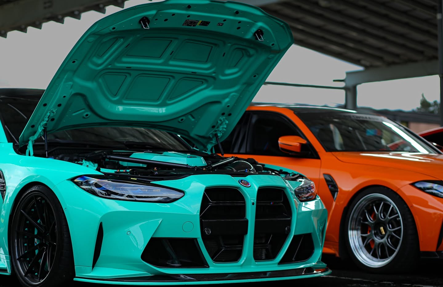 Mint green BMW M4 with its hood open next to an orange M4